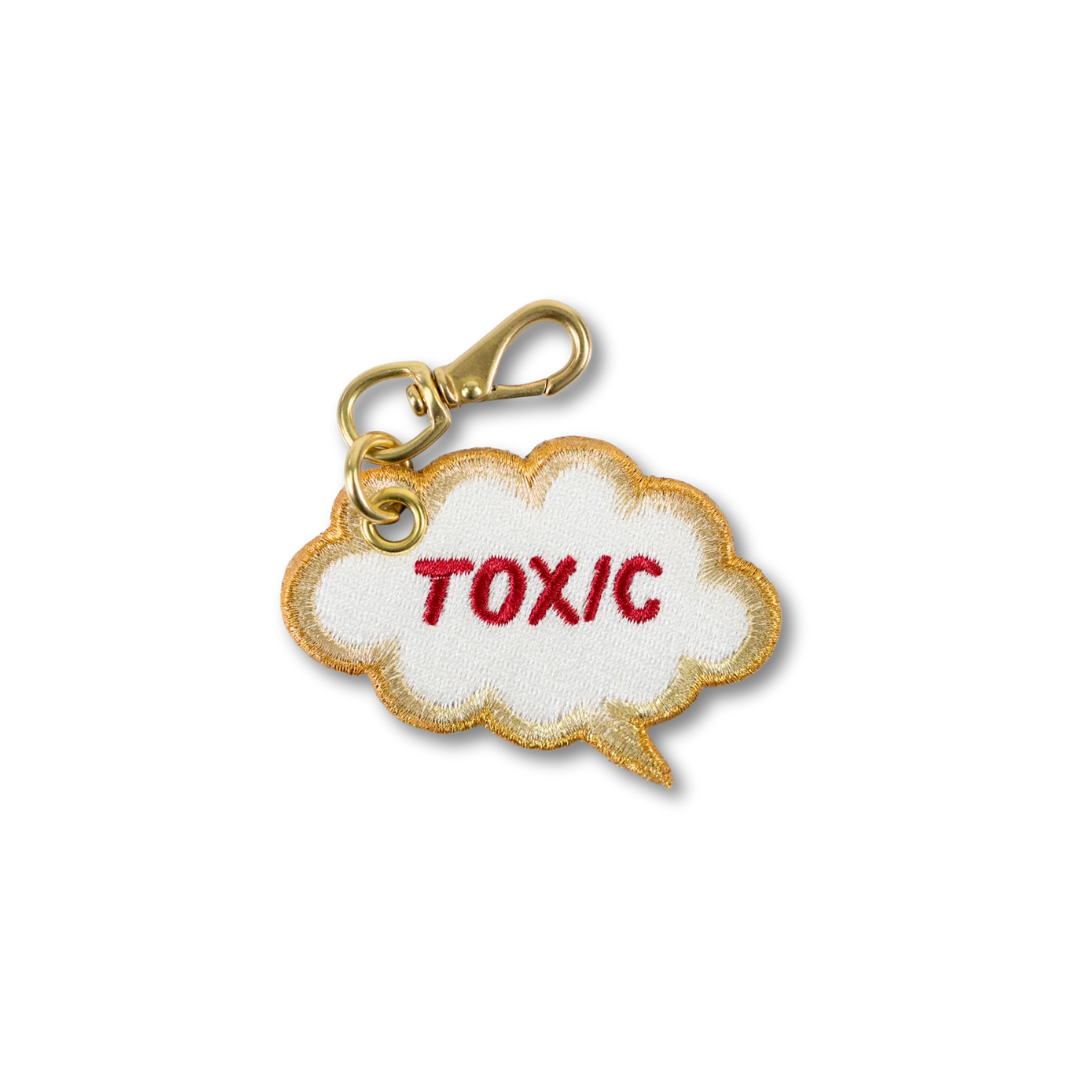 An embroidered cloud-shaped keychain charm with a gold border, white center, and the word 'TOXIC' embroidered in red, attached to a gold lobster clasp on a white background.