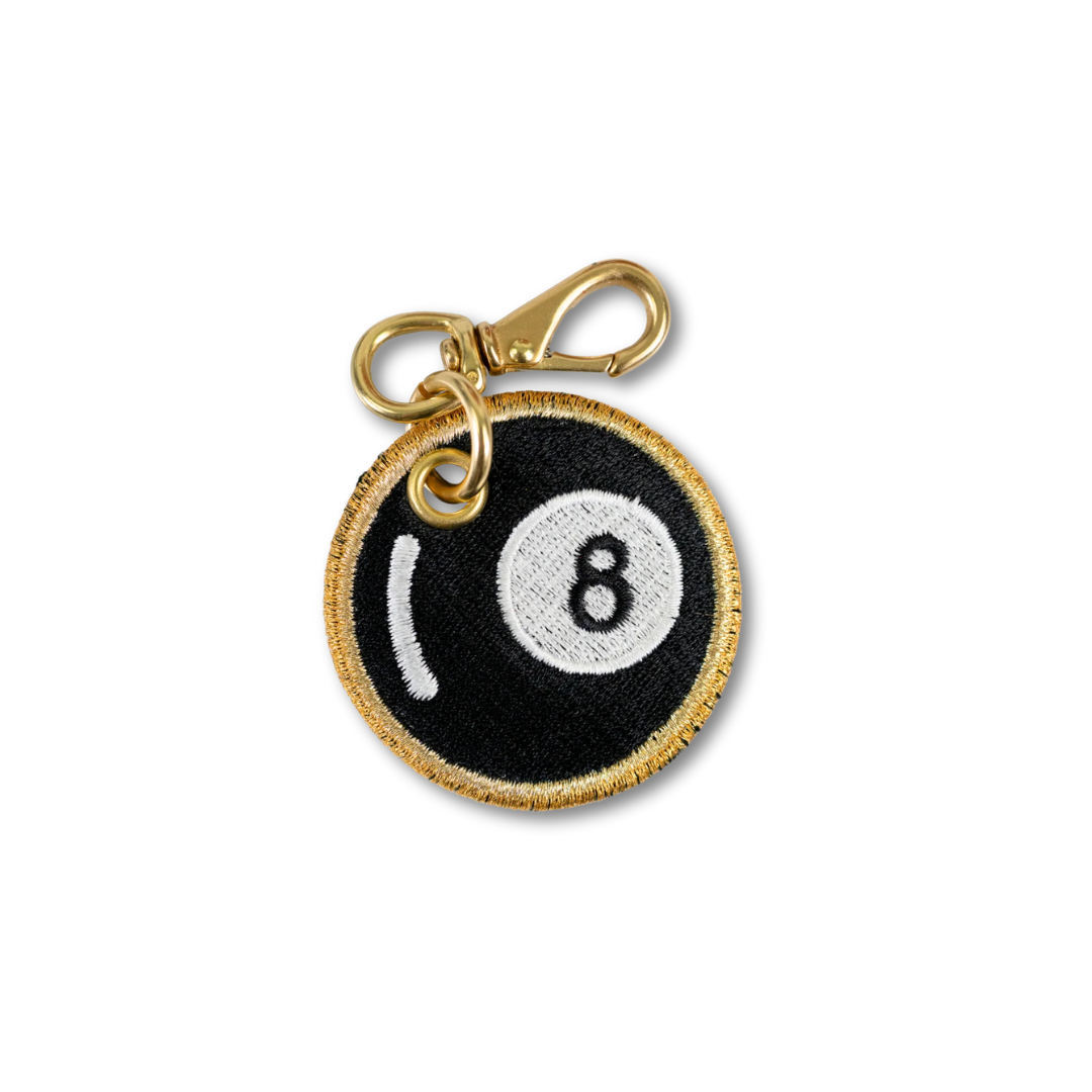An embroidered 8-ball pool cue charm with a gold-tone lobster clasp, featuring a black background, white number 8, and white stripe details with a gold embroidered border.