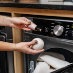 A person holds a small bottle and a white wool ball above a black dryer in a kitchen setting.