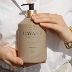 A person in a white shirt holds a bottle of L'Avant Collective Ambre Safran hand soap. The bottle is a warm beige color with a black pump and a gold collar, and the person's hands are visible holding it.