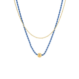 A double-layered necklace featuring a textured blue beaded strand and a fine gold chain, with a small gold bead accent at the center of the blue strand, shown against a plain white background.
