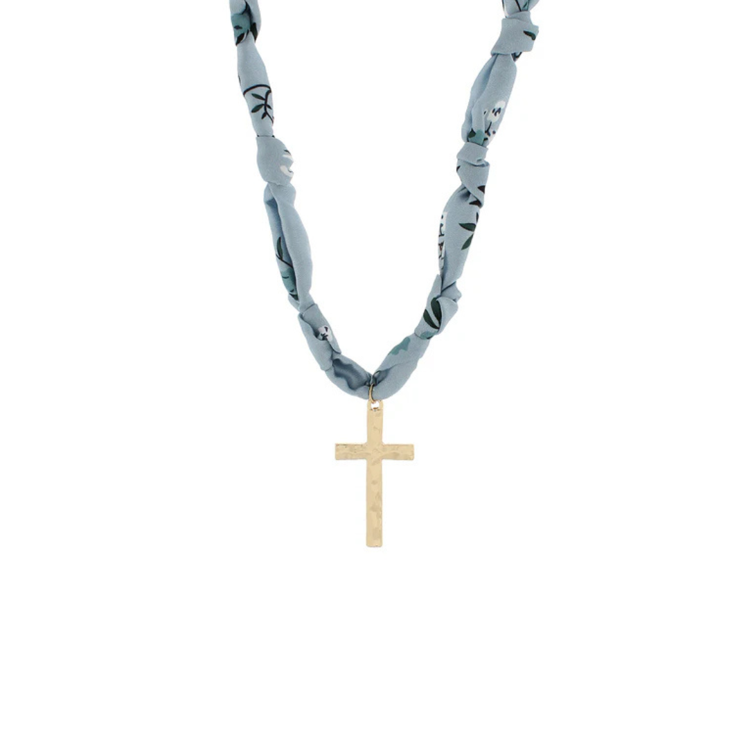 A necklace featuring a light blue and gray patterned fabric chain with a textured gold cross pendant, shown against a plain white background.