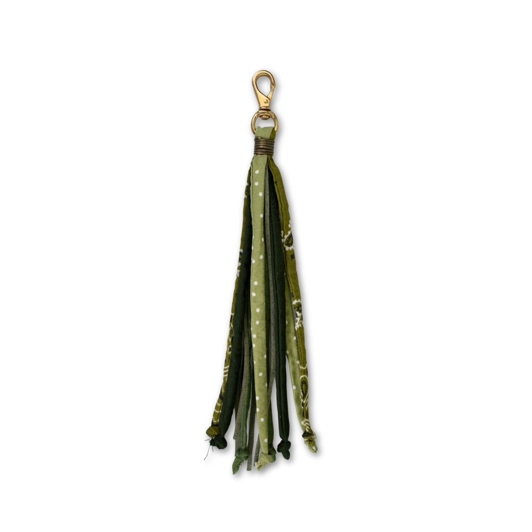 A purse tassel accessory featuring multiple fabric strips in shades of olive green, forest green, and dark green, some with white polka dots and others with a subtle pattern, finished with a gold-tone split ring and lobster clasp.