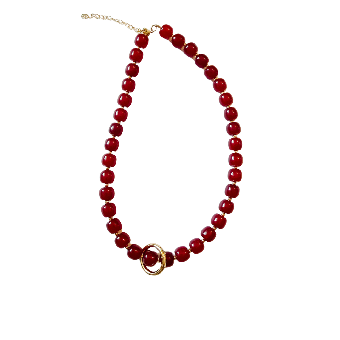 A red beaded necklace with a gold-tone interlocking rings centerpiece and a gold-tone chain extender, displayed on a solid white background.