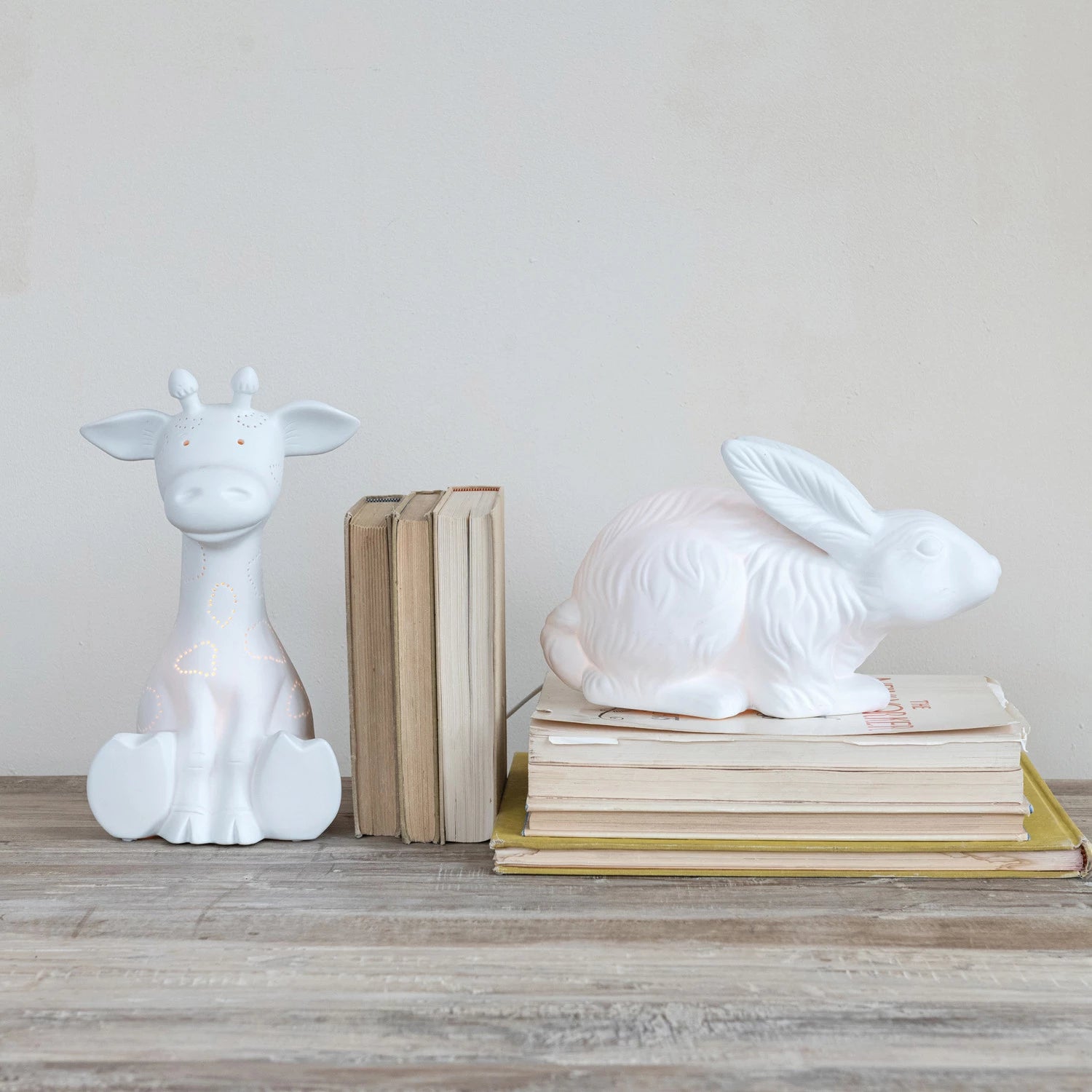 White ceramic giraffe and rabbit lamps on books against a plain wall