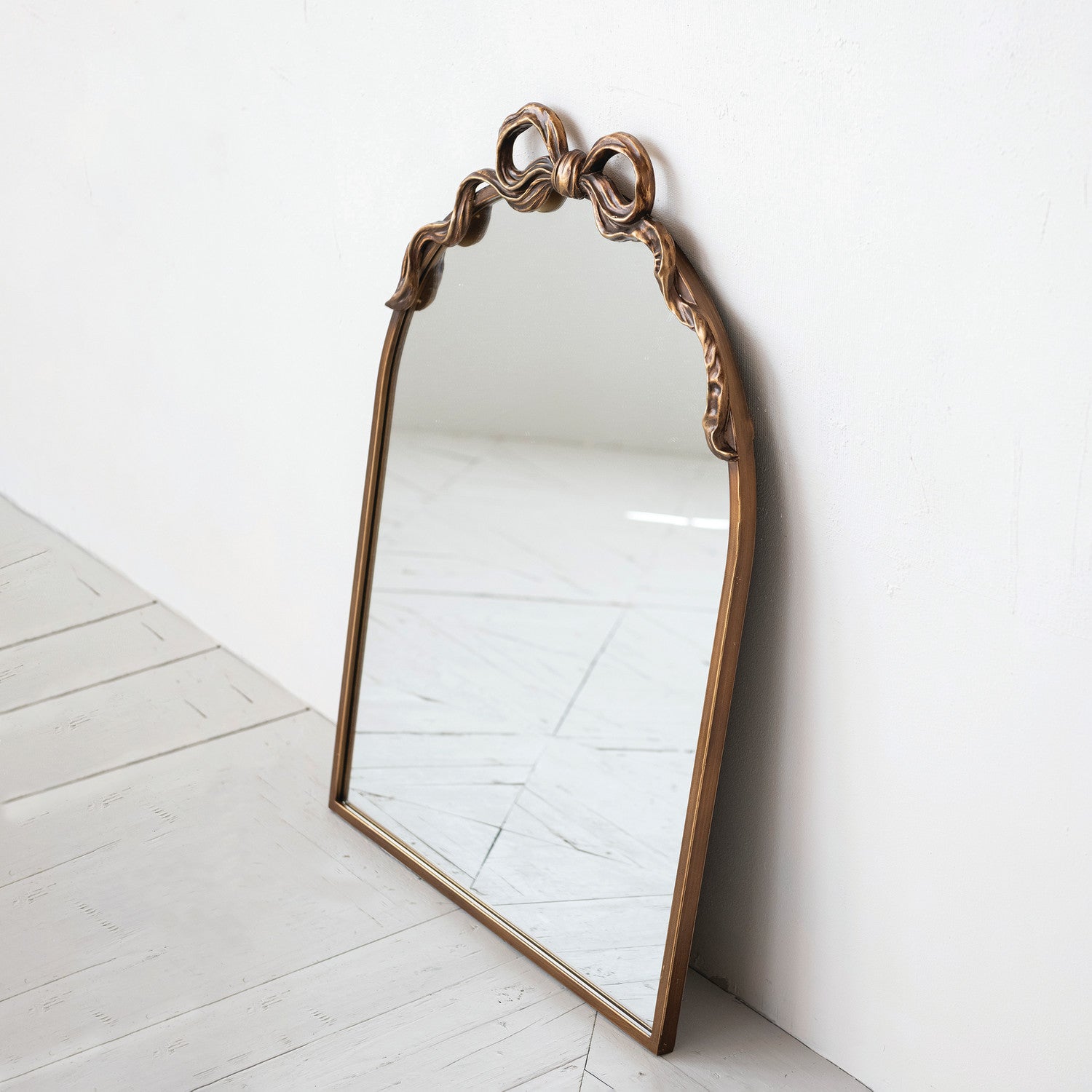 An arched wall mirror with an ornate bronze-finished frame, leaning against a white wall on a light gray wooden floor.