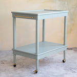A two-tier bar cart in a muted sage green with gold metal hardware, including a handle and casters, set against a textured beige wall and marble floor.