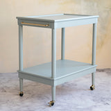 A two-tier bar cart in a muted sage green with gold metal hardware, including a handle and casters, set against a textured beige wall and marble floor.