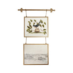 A brass double-hanging wall tapestry with two framed vintage bird and landscape prints suspended from a brass rod against a white background.