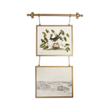 A brass double-hanging wall tapestry with two framed vintage bird and landscape prints suspended from a brass rod against a white background.
