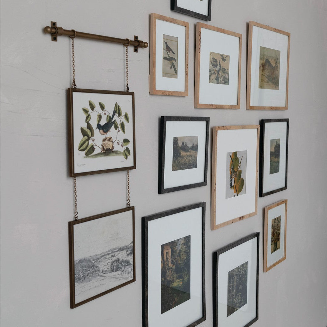 A gallery wall arrangement of multiple framed botanical and landscape prints on a light gray wall, including one hanging print suspended from a horizontal rod.