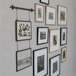 A gallery wall arrangement of multiple framed botanical and landscape prints on a light gray wall, including one hanging print suspended from a horizontal rod.
