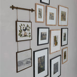 A gallery wall arrangement of multiple framed botanical and landscape prints on a light gray wall, including one hanging print suspended from a horizontal rod.