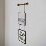 A rustic wall-mounted hanging rod with two framed art prints suspended by chains against a white textured wall.
