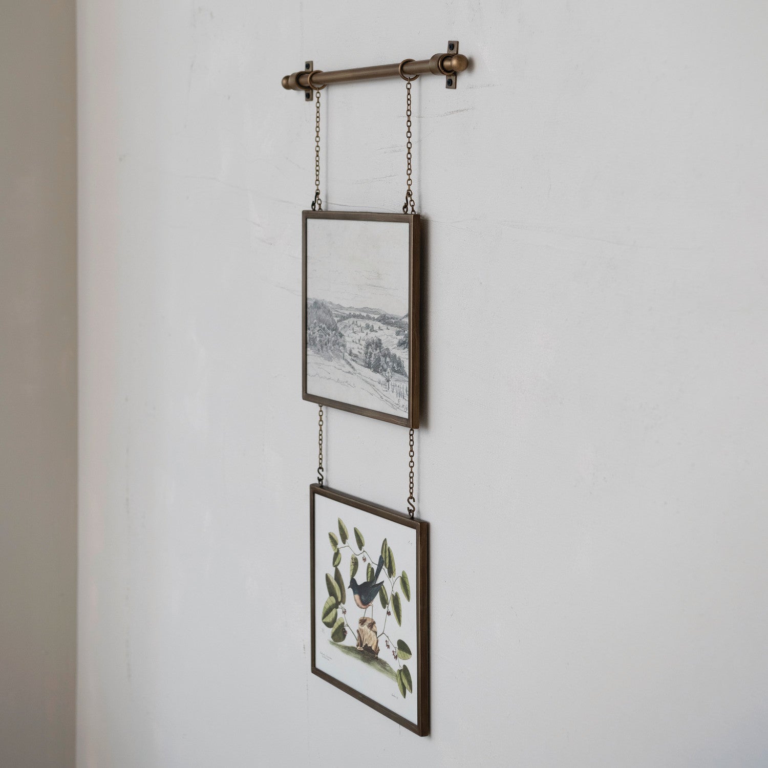 A rustic wall-mounted hanging rod with two framed art prints suspended by chains against a white textured wall.