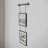 A rustic wall-mounted hanging rod with two framed art prints suspended by chains against a white textured wall.