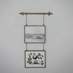 Two framed prints hanging on a wall with a decorative metal rod and chains.