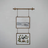Two framed prints hanging on a wall with a decorative metal rod and chains.