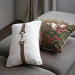 Two decorative pillows on a gray sofa, one with a brown strap and white background, the other with a nature-themed design.