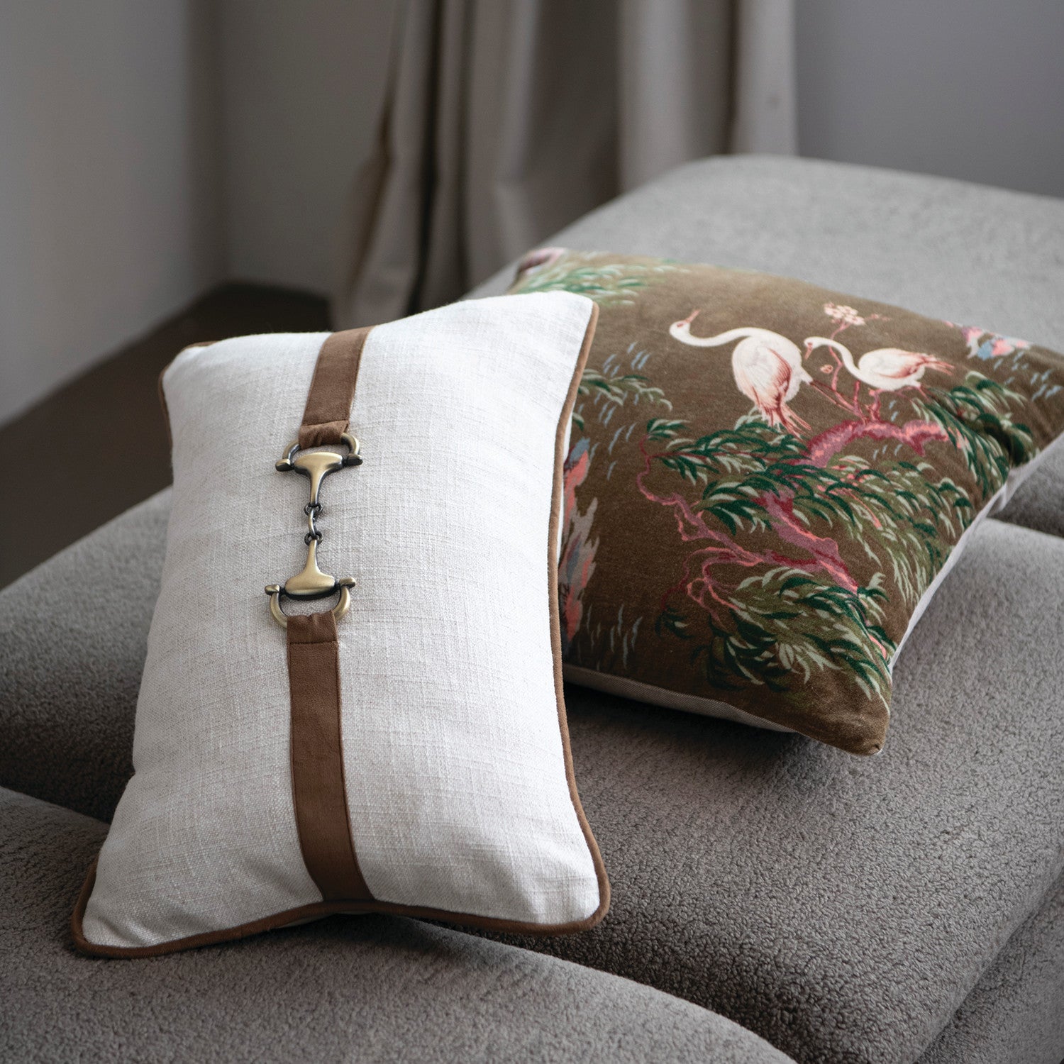 Two decorative pillows on a gray sofa, one with a brown strap and white background, the other with a nature-themed design.