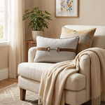 A cozy living room scene featuring a cream upholstered armchair with a beige throw blanket draped over it, a matching accent pillow with a brown leather-look strap and horsebit detail, and a plaid throw pillow. A small wooden side table with a potted plant and framed wall art are visible in the background.