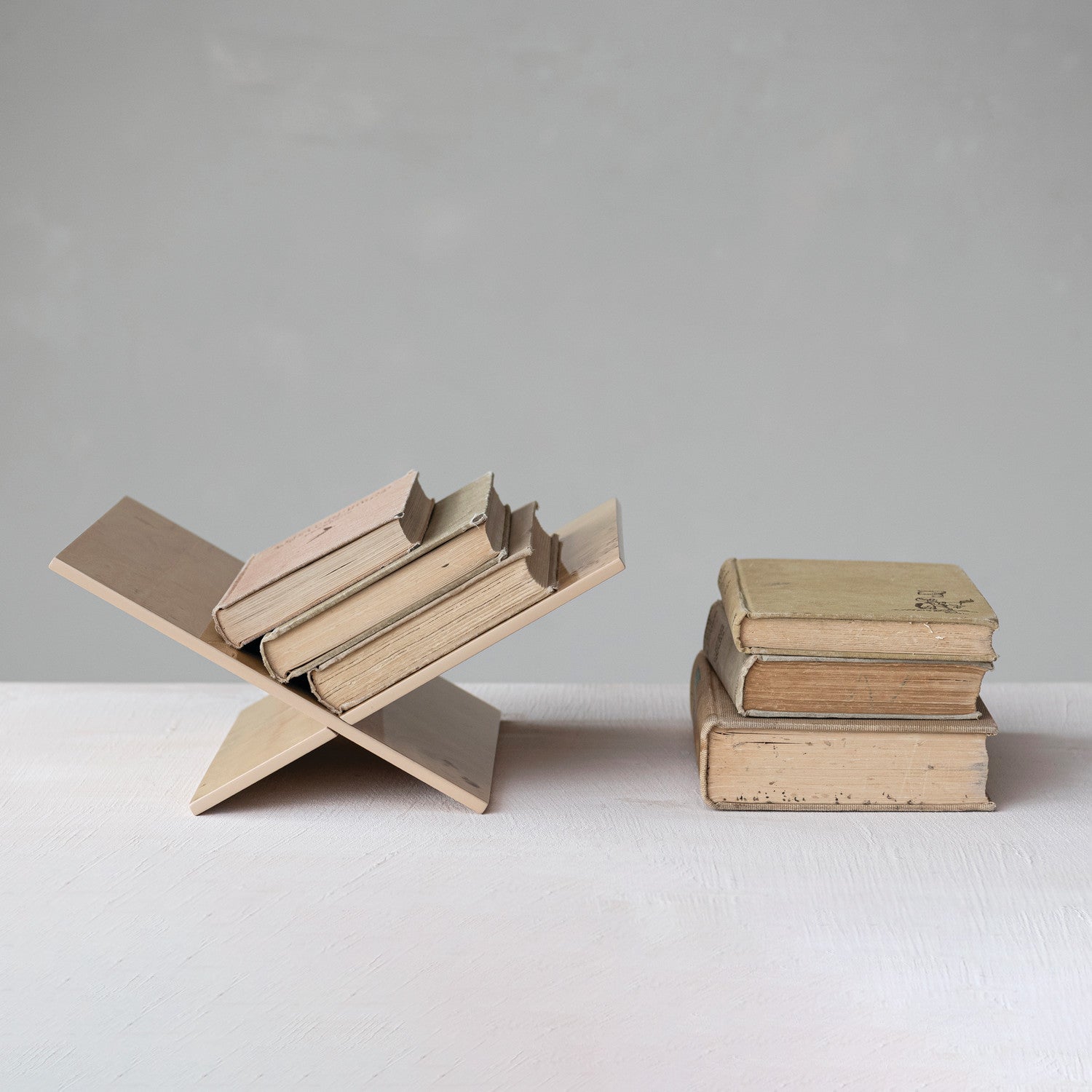 A minimalist wooden bookshelf sculpture holding several vintage books, displayed next to a stack of three old books on a white surface against a neutral gray background.