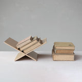 A minimalist wooden bookshelf sculpture holding several vintage books, displayed next to a stack of three old books on a white surface against a neutral gray background.