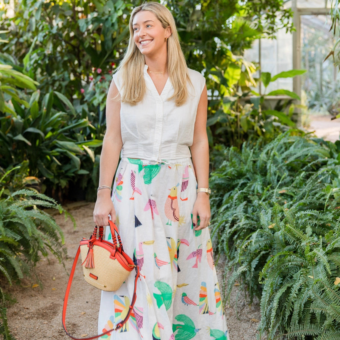 Woman in a white blouse and colorful skirt standing in a greenhouse holding a Montemare Palma Crossbody with orange leather accents