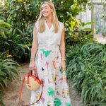 Woman in a white blouse and colorful skirt standing in a greenhouse holding a Montemare Palma Crossbody with orange leather accents