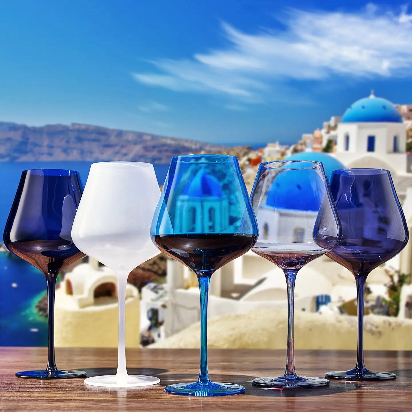 Five colored wine glasses on a table with a scenic background of blue-domed churches.
