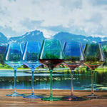 Five colorful wine glasses with different colored liquids on a wooden table with a scenic background of mountains and water.