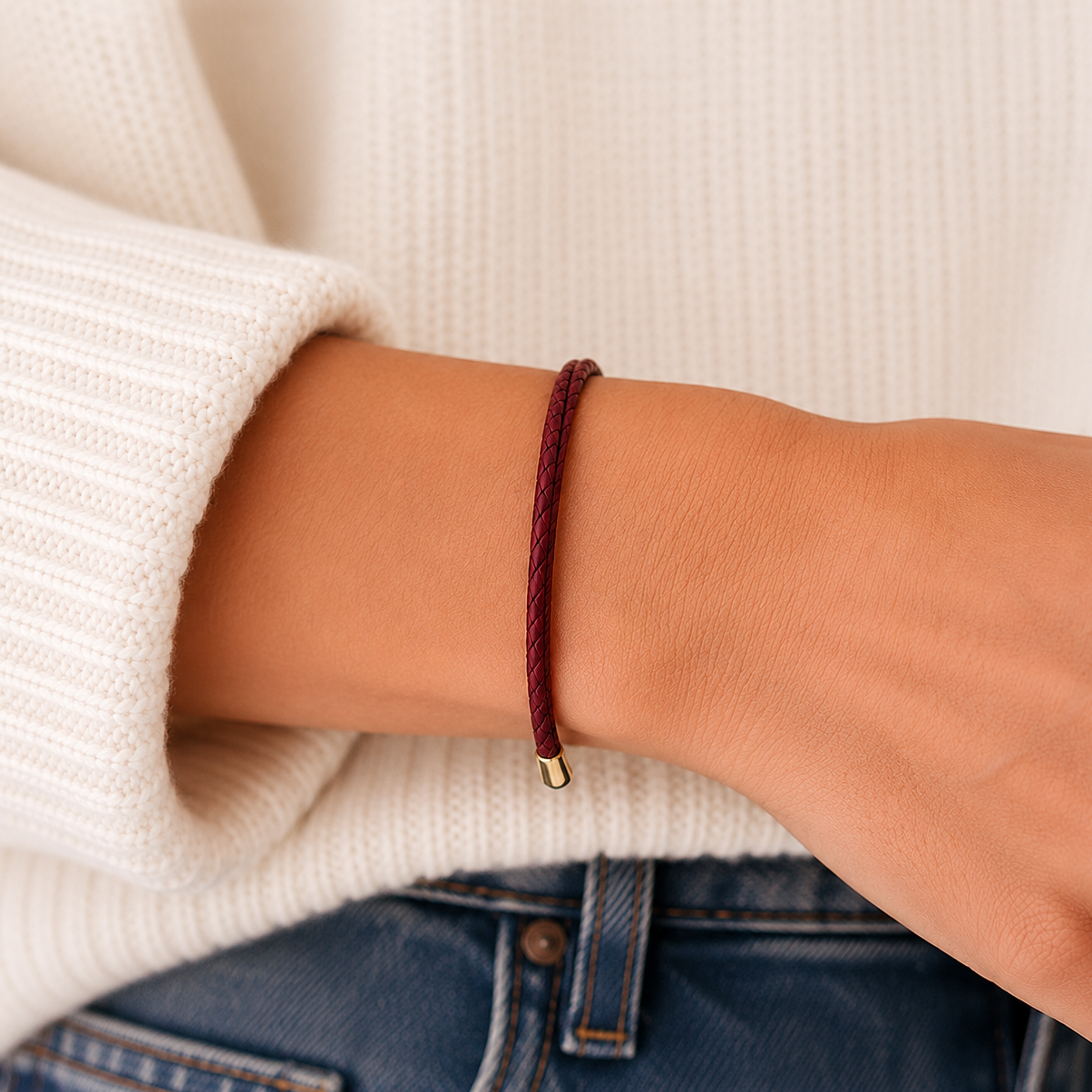A close-up of a person's wrist wearing a thin braided cord bracelet in burgundy with a small gold bead accent, styled with a cream ribbed knit sweater and blue jeans.