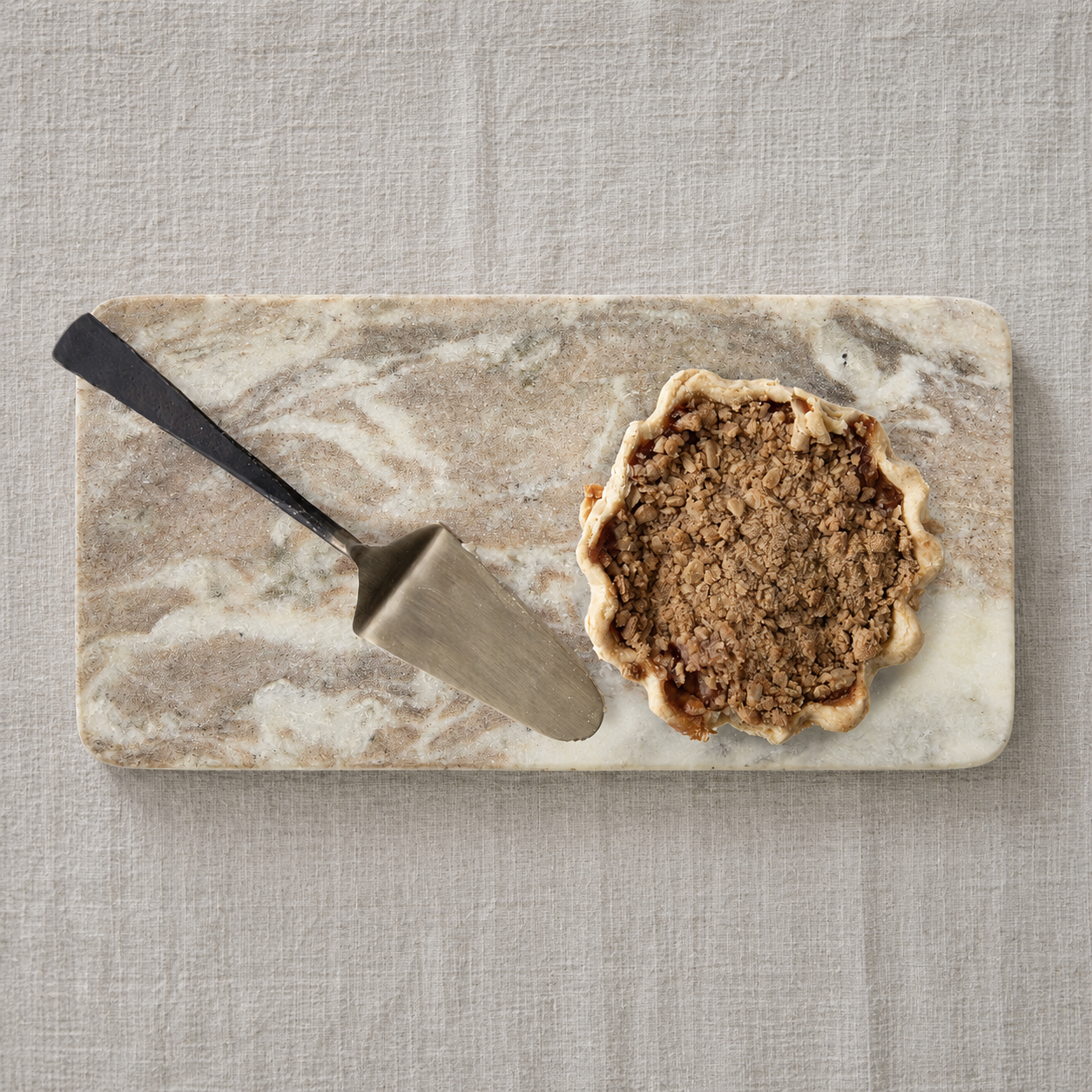 A small scalloped pie with a crumb streusel topping sits on a rectangular marble serving board with a metal pie server beside it, all placed on a light beige linen surface.