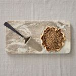 A small scalloped pie with a crumb streusel topping sits on a rectangular marble serving board with a metal pie server beside it, all placed on a light beige linen surface.