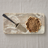 A small scalloped pie with a crumb streusel topping sits on a rectangular marble serving board with a metal pie server beside it, all placed on a light beige linen surface.