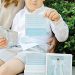 Child holding a prayer card with text, sitting next to an adult outdoors.