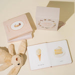 Children's book with illustrations of a teddy bear, ice cream cone, and cake on a beige surface.