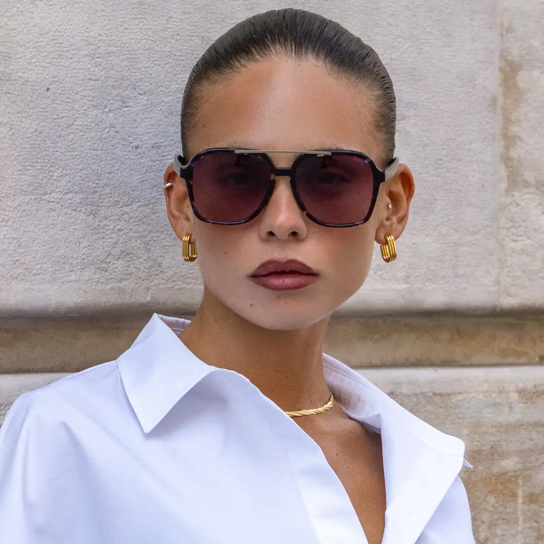 Woman wearing sunglasses and a white shirt against a beige wall.