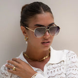 Woman wearing gold sunglasses, earrings, and jewelry against a neutral background