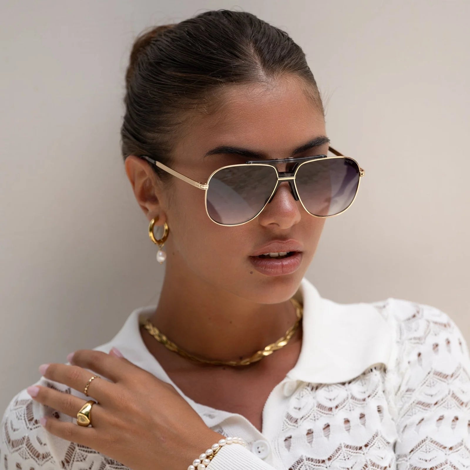 Woman wearing gold sunglasses, earrings, and jewelry against a neutral background