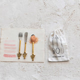Gold cutlery set with a salt shaker on a textured surface, accompanied by a branded drawstring bag.