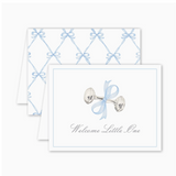 Folded greeting card with a watercolor-style silver baby rattle tied with a blue bow and the text “Welcome Little One” on the front; back features a blue bow lattice pattern. Card is blank inside and includes a Euro flap envelope.