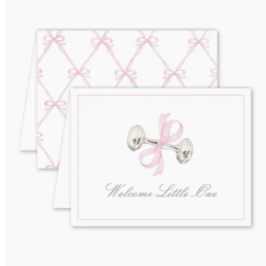 Folded greeting card with a watercolor-style silver baby rattle tied with a pink bow and the text “Welcome Little One” on the front. Card is blank inside and includes a Euro flap envelope.