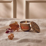 Candle with a wooden lid on a white fabric surface with autumn leaves.