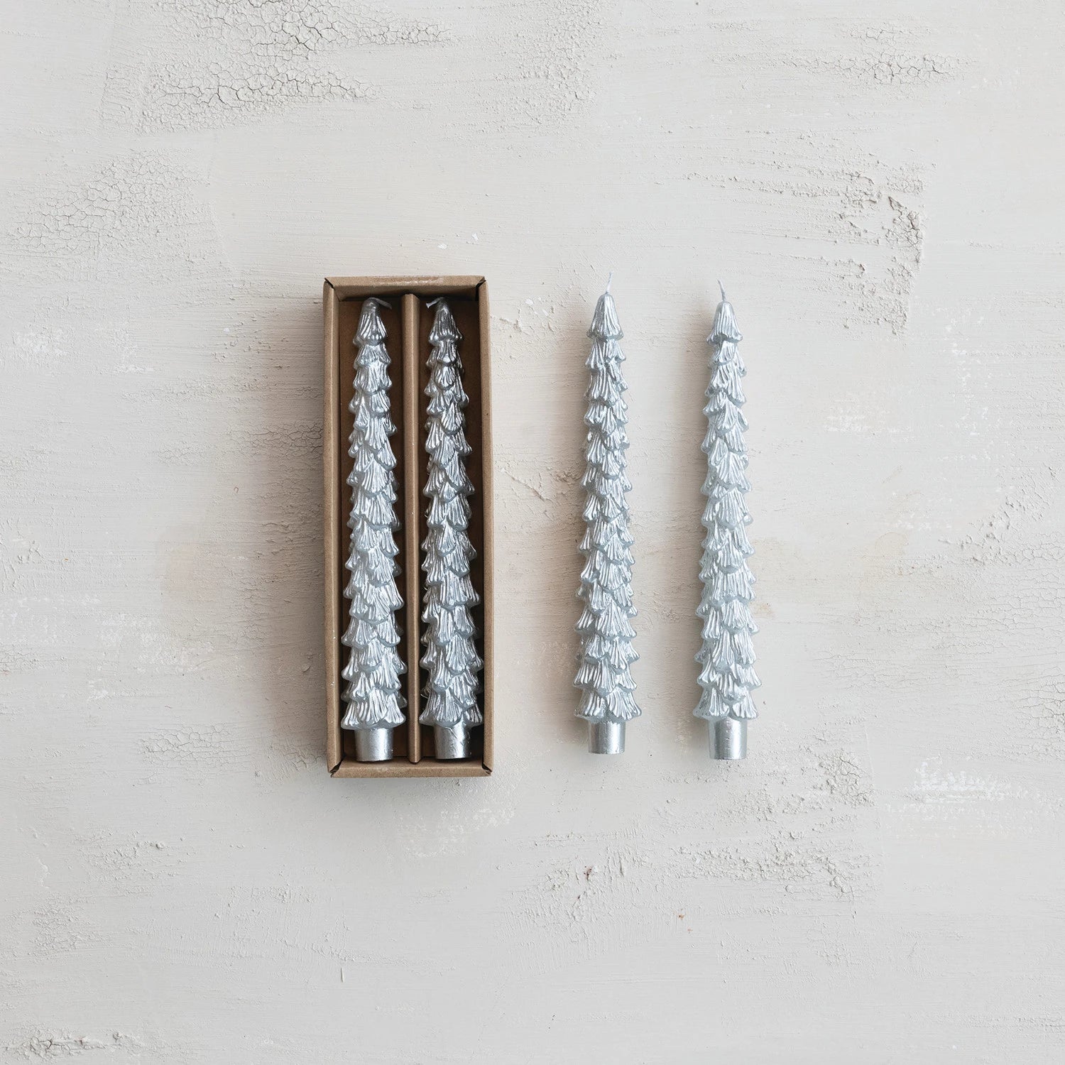 Two silver Christmas tree-shaped candles on a textured white surface with one candle in a box.