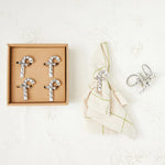 Silver cross-shaped napkin holders in a cardboard box on a textured white surface.